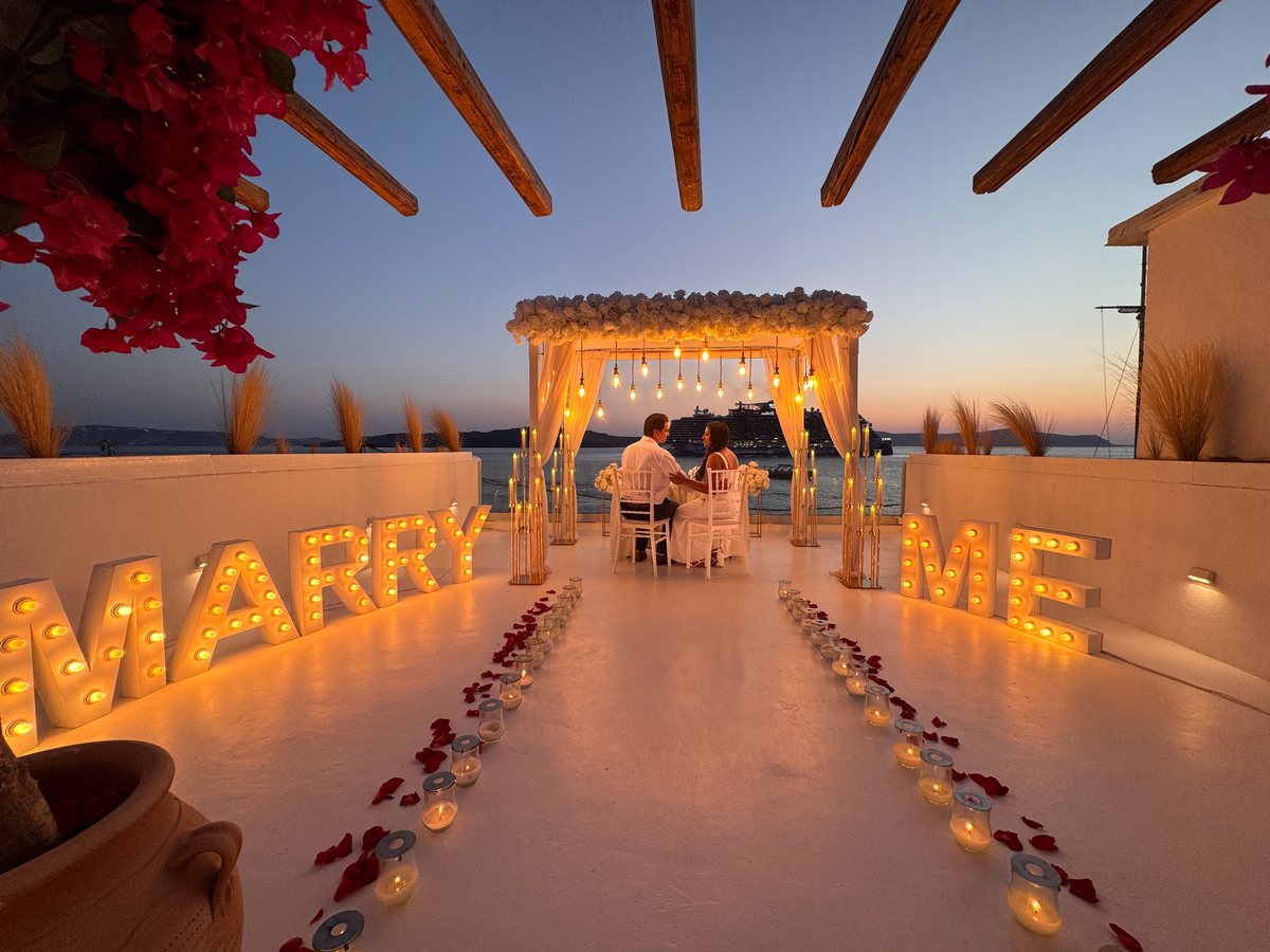 MARRY ME in lit letters on AION's terrace at dusk — a candle and petal aisle leading to the white canopy