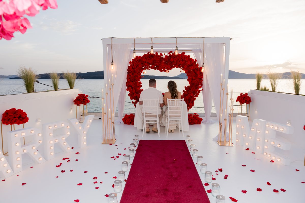 A couple at their table on AION's terrace, MARRY ME letters and the red rose heart beside them
