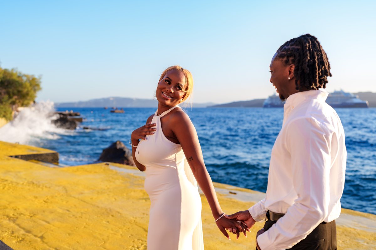 A couple at the old port breakwater beside AION in full daylight, the caldera across the water