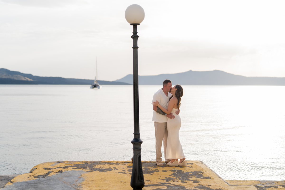 A couple kissing at the end of the old port pier at AION, the caldera behind them