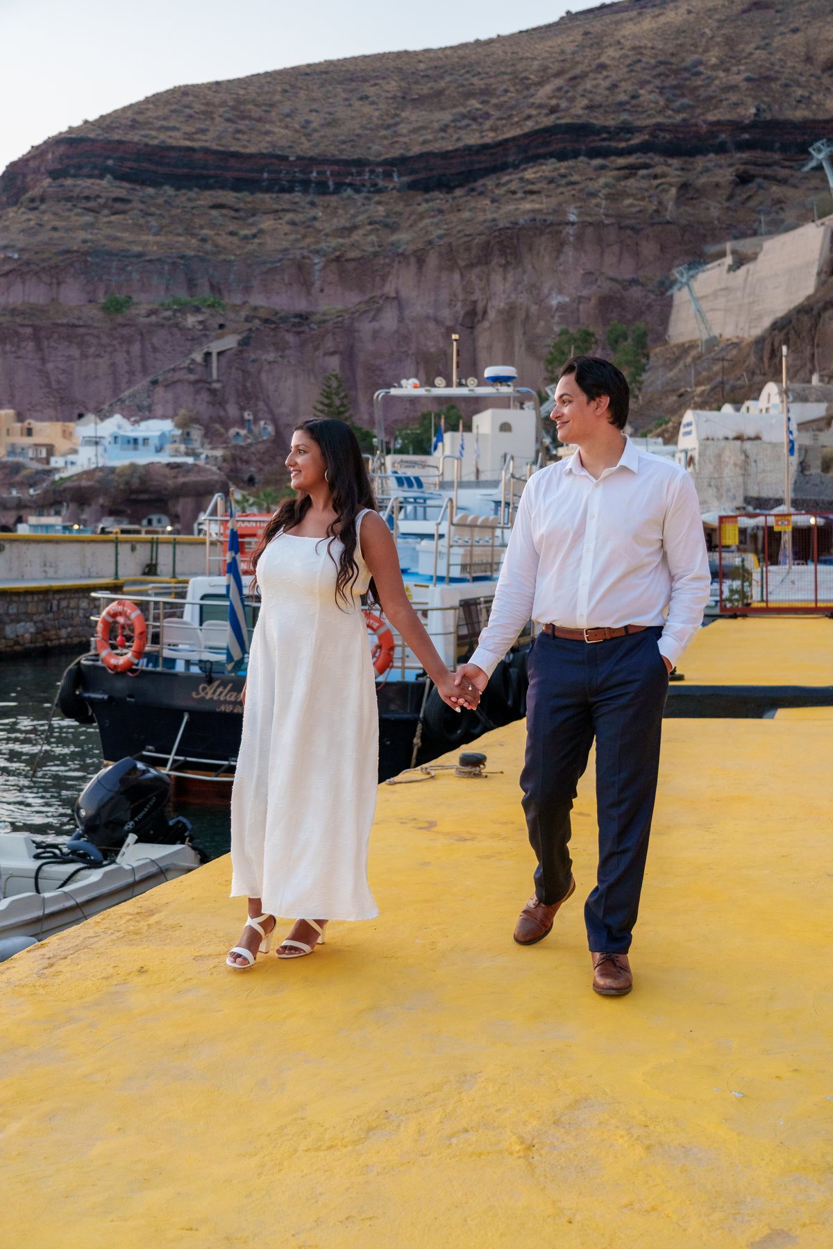 A couple walking AION's old port quay in the evening, fishing boats and the Fira cliff behind