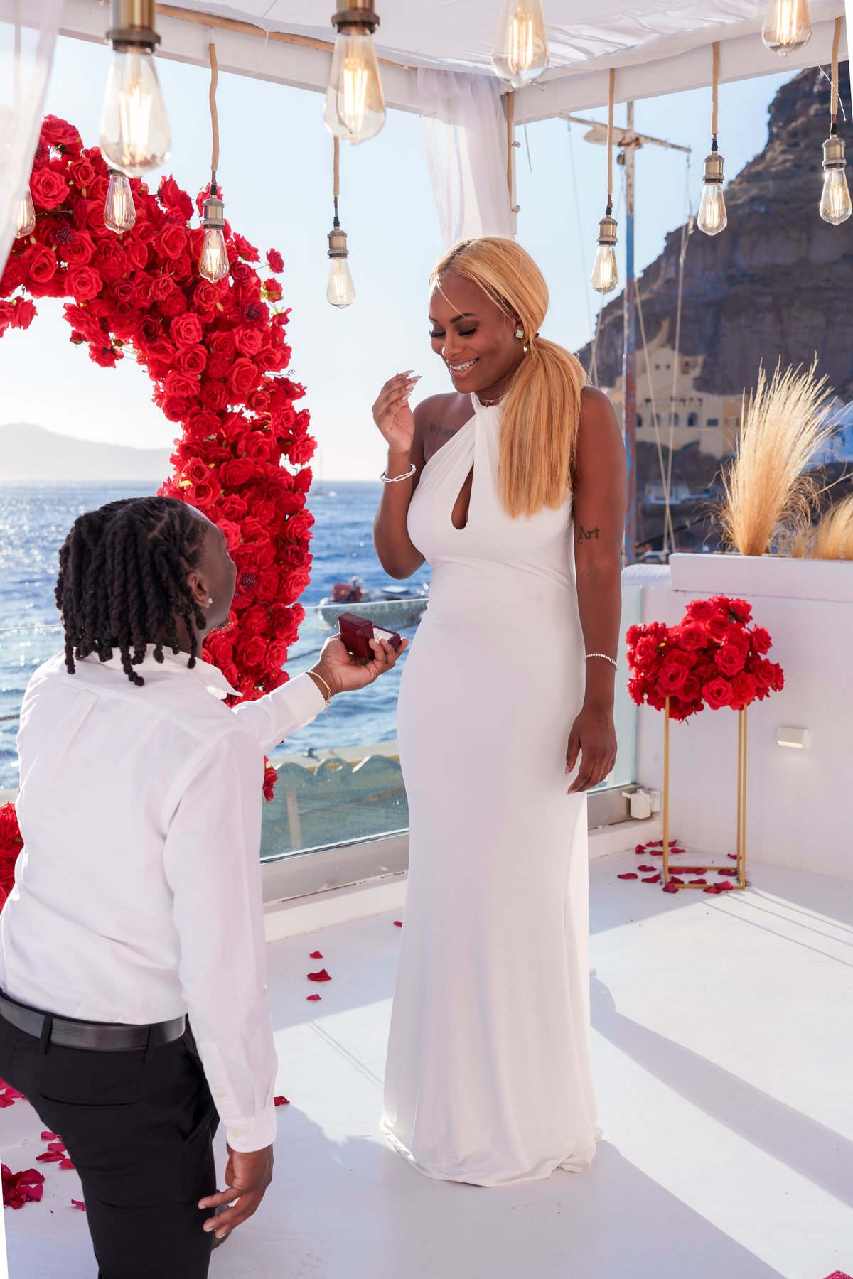 A proposal on one knee under the red rose heart on AION's terrace, blue sea behind