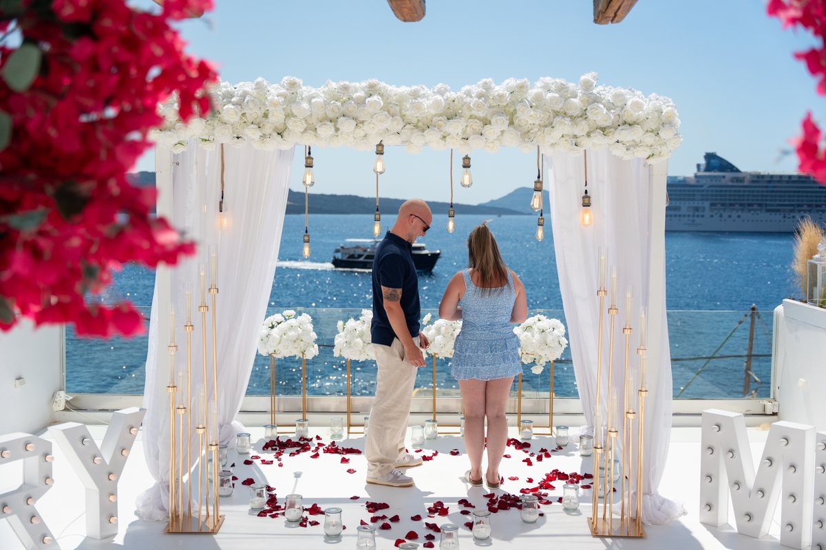 A couple beside the bougainvillea at AION's arch, a cruise ship on the caldera beyond