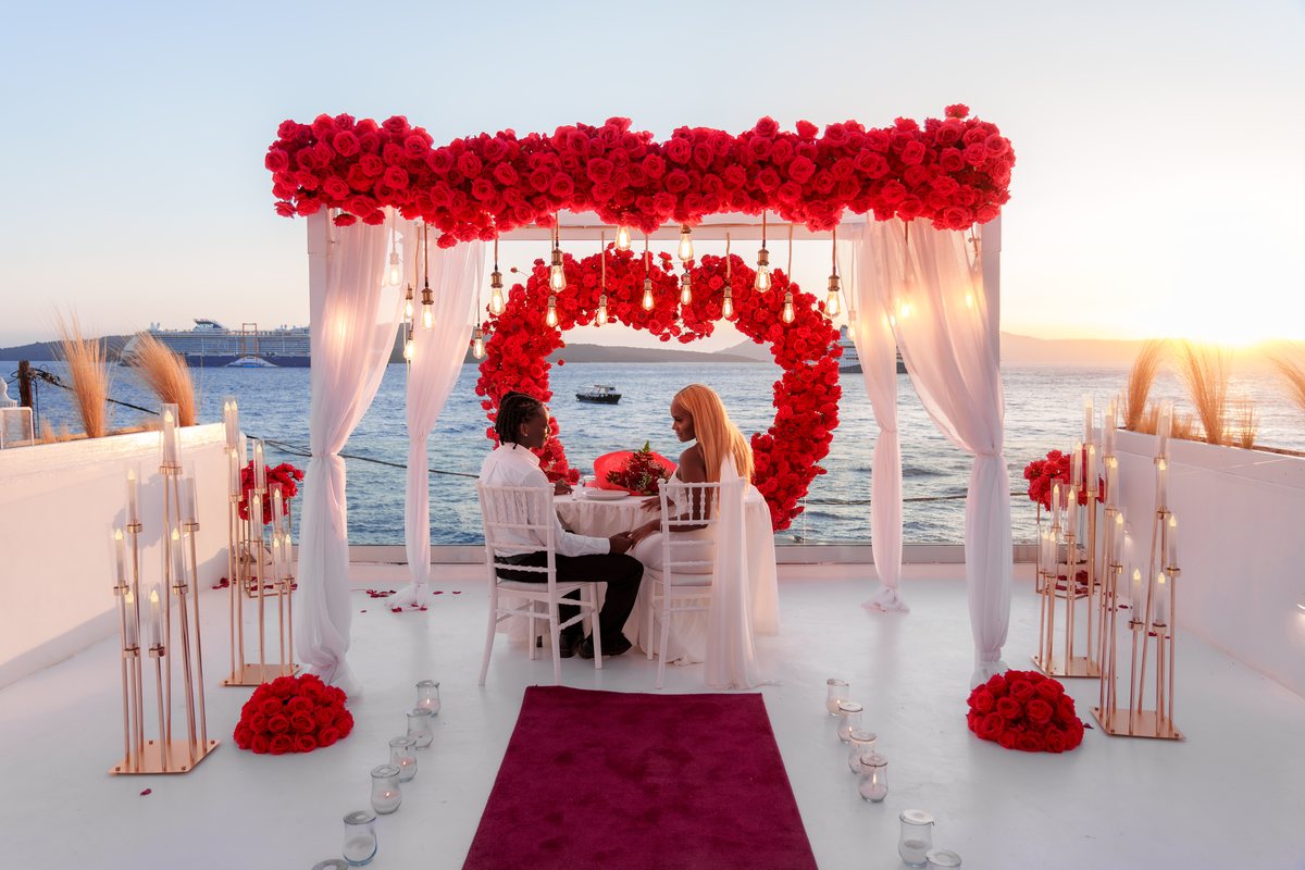 A couple at their dinner table under AION's red rose canopy as the sun sets over the caldera