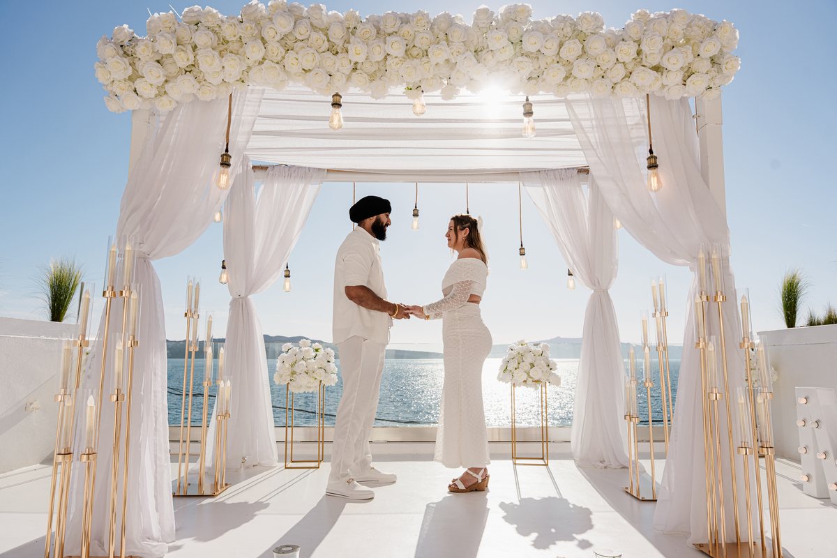A wedding couple hand in hand under AION's white flower arch, the caldera wide open behind them