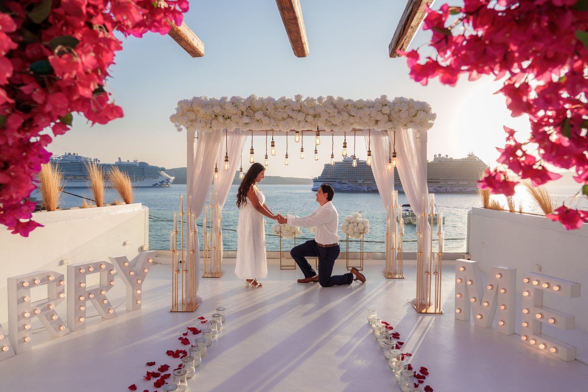 A proposal on one knee under AION's white flower canopy, bougainvillea overhead and a cruise ship on the caldera