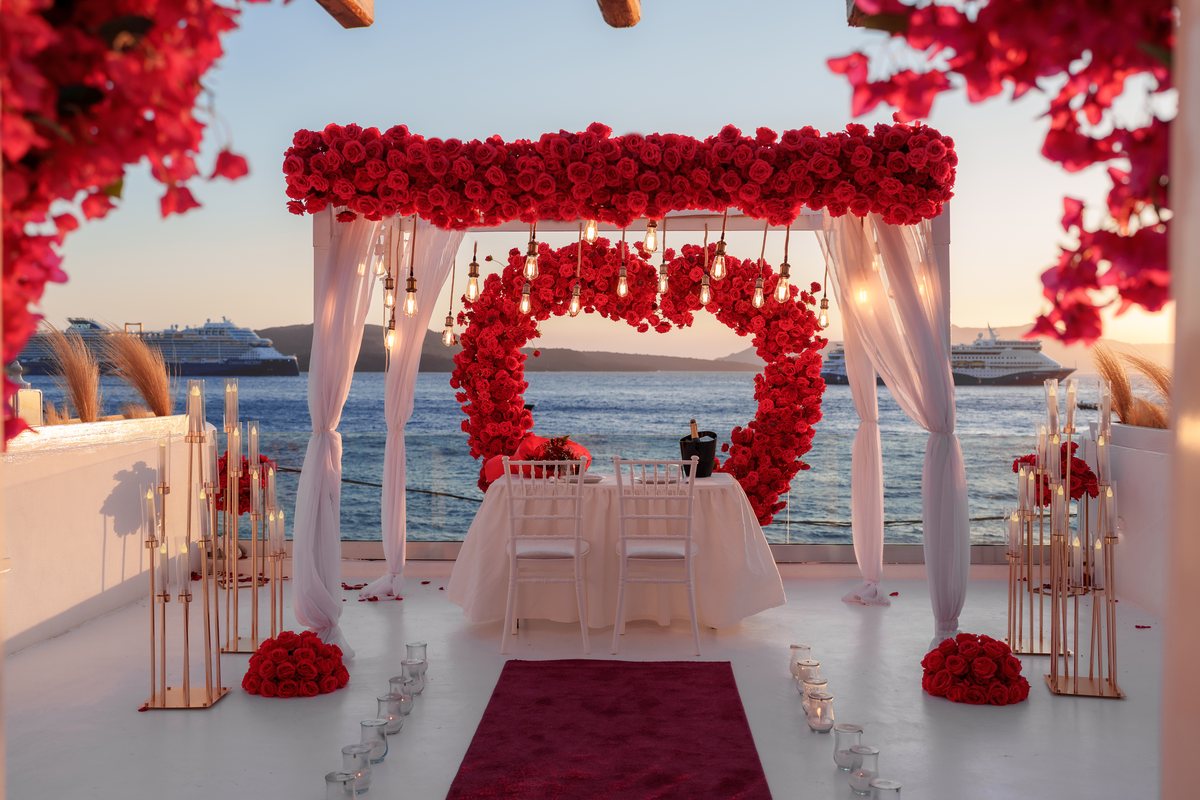 Sunset from AION's terrace — the red rose canopy, heart wreath and a table for two facing the caldera