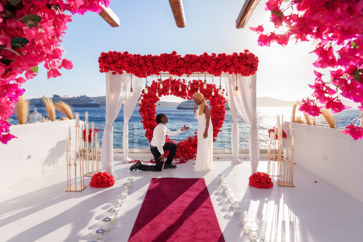 The proposal moment at AION Santorini — one knee before the red rose heart, bougainvillea overhead and the caldera behind