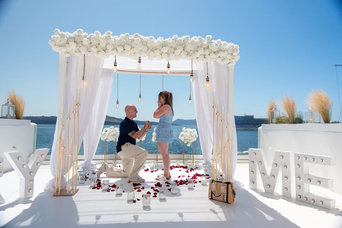 A surprise proposal on one knee under AION's white arch, rose petals across the terrace