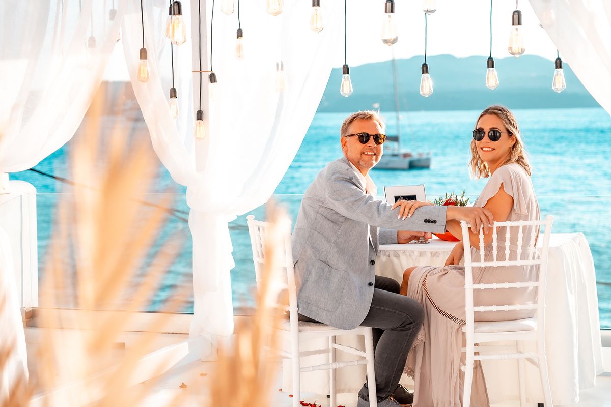 Scot and his fiancee at the table under AION's bulbs and curtains, a catamaran on the caldera behind