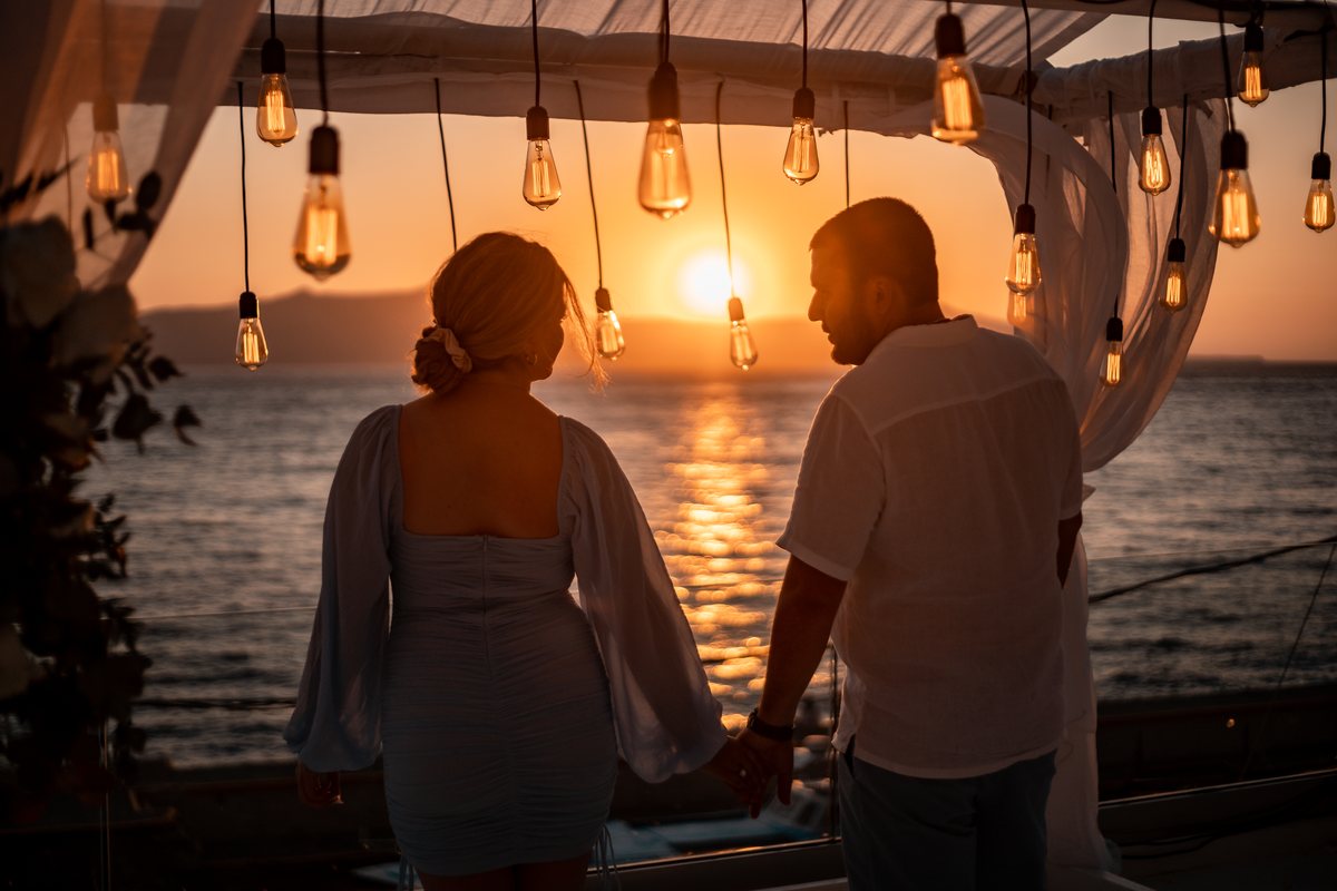 The Santorini sunset from AION's terrace, a couple in silhouette beneath the hanging bulbs