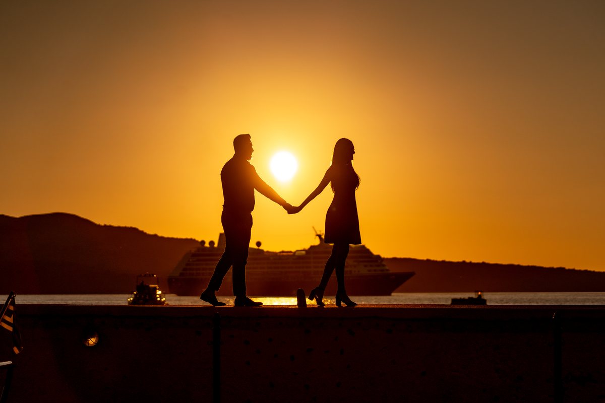 A couple holding hands in silhouette as the sun drops into the sea below AION