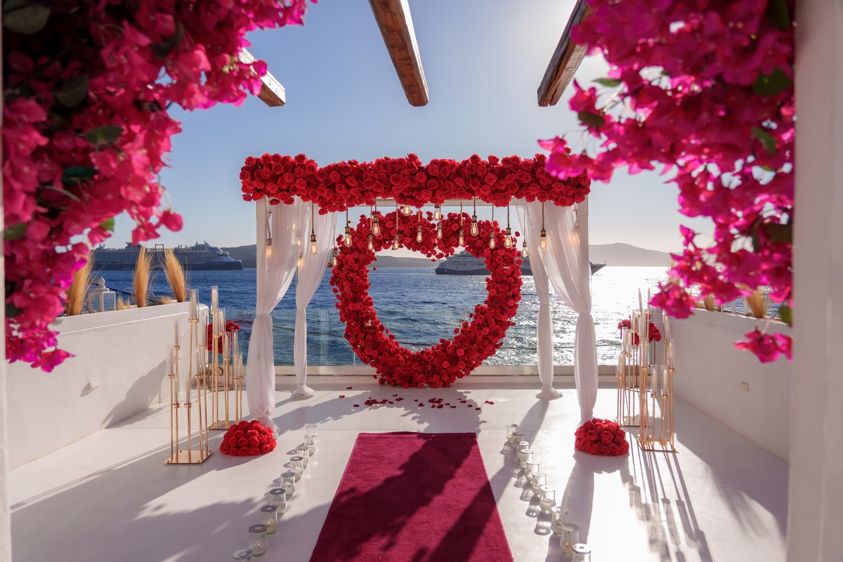 The heart-shaped red rose arch on AION's terrace, bougainvillea overhead and the caldera behind